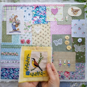 May include: A handmade patchwork quilt with various fabric squares. The quilt features floral patterns, embroidered images of a bunny, bird, and butterflies, and decorative embellishments. The color palette includes pastels and earth tones.