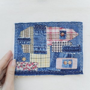 Può includere: Un'opera d'arte tessile rettangolare con un design patchwork. La base è in denim blu, con varie toppe di tessuto a motivi scozzesi, floreali e a righe. Le toppe sono fissate con cuciture bianche visibili e decorate con piccoli bottoni rotondi.