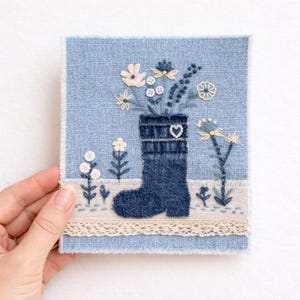 May include: Embroidered denim art piece showing a blue denim boot filled with embroidered flowers in white and blue. The artwork is on a light blue denim background with a cream lace trim. The boot has a heart detail.