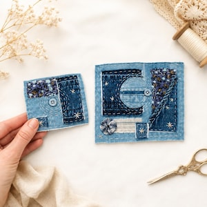 May include: Two denim fabric art pieces with a celestial theme. The pieces feature patches of blue denim, floral fabric, and white stitching. One piece is square, the other is rectangular. Both have button and star accents. A hand holds one piece.
