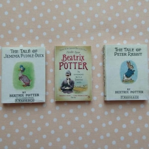 Può includere: Tre libri in miniatura di Beatrix Potter su uno sfondo a pois. I titoli includono "The Tale of Jemima Puddle-Duck" e "The Tale of Peter Rabbit". Le copertine presentano illustrazioni di un'anatra e di un coniglio.