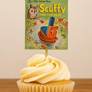 Może przedstawiać: Ż&oacute;łty cupcake z białym kremem, ozdobiony małą złotą książeczką przedstawiającą holownik o imieniu "Scuffy" z tekstem "The Tug Boat".