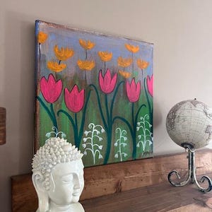 May include: A painting of a field of pink and yellow flowers on a canvas. The painting is on a wooden shelf with a white Buddha statue and a globe.