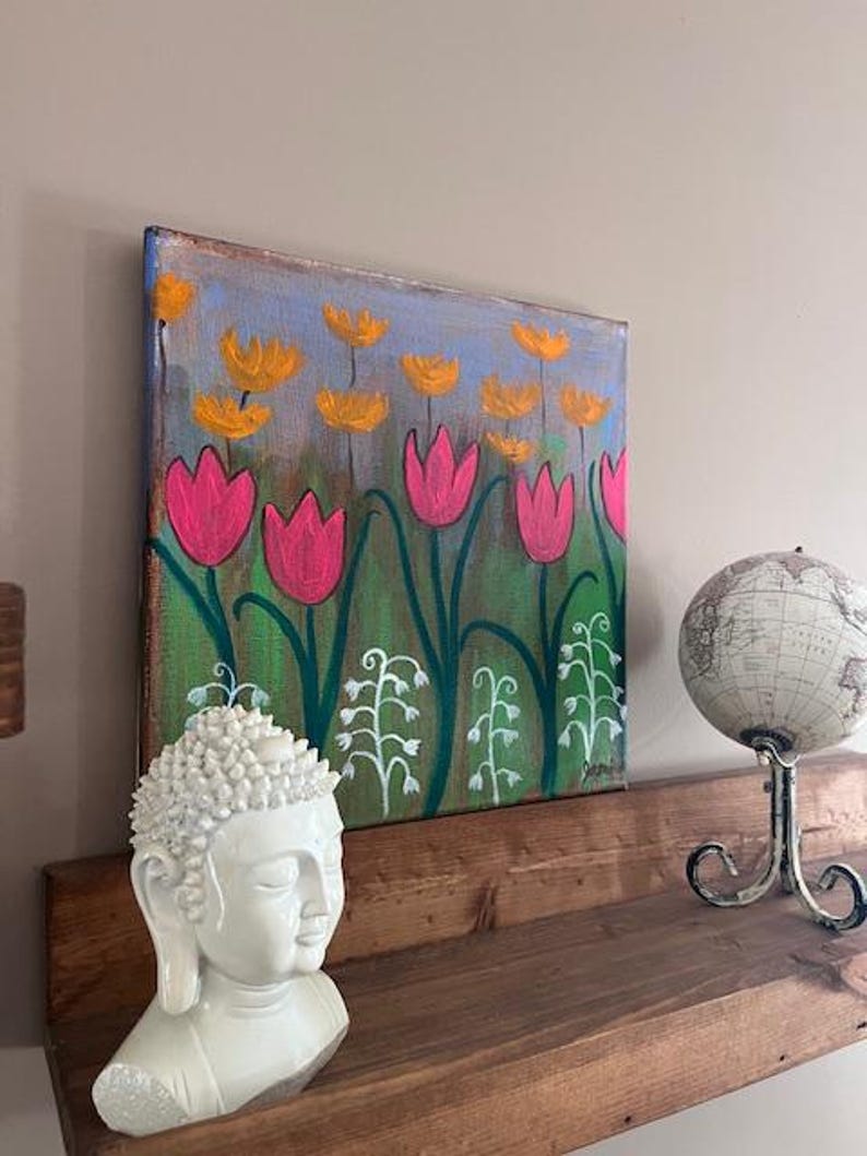 May include: A painting of a field of pink and yellow flowers on a canvas. The painting is on a wooden shelf with a white Buddha statue and a globe.