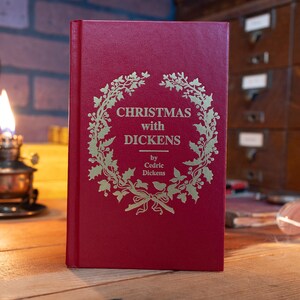Pode incluir: Um livro de capa dura vermelho intitulado Christmas with Dickens por Cedric Dickens, apresentando uma ilustração de uma coroa dourada na capa frontal. O livro está posicionado na vertical, exibindo o título claramente no centro da coroa.