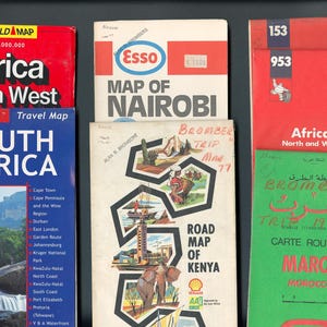 May include: A collection of vintage travel maps including Africa North West, Map of Nairobi, South Africa, Road Map of Kenya, and Maroc. The maps feature various colors, illustrations, and text, with some covers showing handwritten notes and dates.