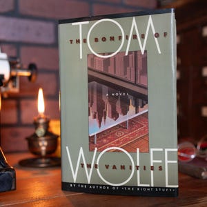 May include: A hardcover book titled The Bonfire of the Vanities by Tom Wolfe with a sage green cover. The front features a central illustration of a city skyline reflected in a glass surface above a patterned rug, with white text throughout the cover.