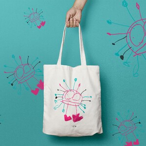 Puede incluir: Una bolsa de tela blanca con un diseño colorido dibujado a mano de una criatura con dos ojos y pies rosas. El diseño se repite en el fondo azul detrás de la bolsa.