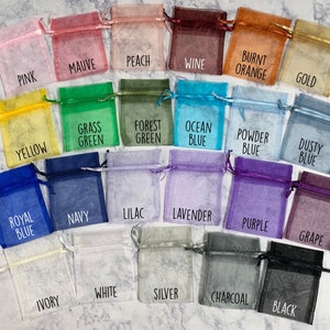 May include: A collection of 20 small, sheer fabric bags in various colors, including pink, mauve, peach, wine, burnt orange, gold, yellow, grass green, forest green, ocean blue, powder blue, dusty blue, royal blue, navy, lilac, lavender, purple, grape, ivory, white, silver, charcoal, and black. Each bag has a label with its corresponding color name.