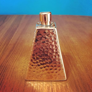May include: A hammered copper bottle with a silver top. The bottle is shaped like a pyramid and has a textured surface.