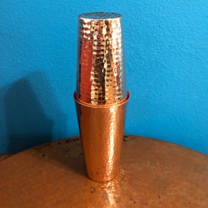 May include: A copper cocktail shaker with a hammered texture. The shaker consists of two parts: a larger base and a smaller, inverted top. The copper has a warm, reddish-brown hue, and the hammered finish adds visual interest. The background is a solid blue.