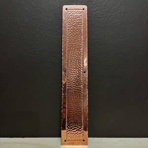 May include: A copper-colored, rectangular door pull with a hammered texture. The door pull has a series of small, evenly spaced holes along the edges and a central dotted pattern. The metal has a warm, reddish-brown hue.