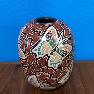 May include: A ceramic vase with a black rim and a rounded body. The vase features a repeating pattern of butterflies and abstract shapes in shades of red, cream, and black. The butterflies have green, yellow, and blue accents.