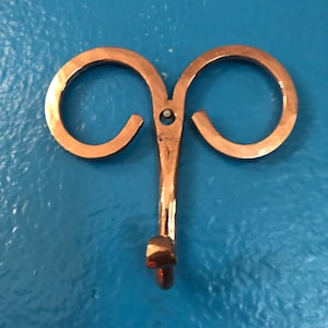 May include: A copper-colored metal wall hook with a unique design. The hook features two curved loops at the top, connected to a central stem that ends in a hook. The hook is mounted on a blue surface.