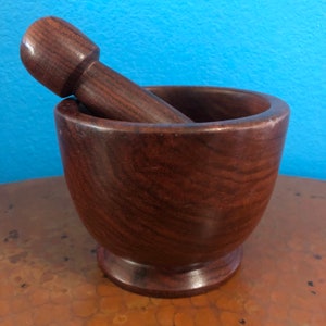 May include: A dark brown wooden mortar and pestle set. The mortar is a bowl shape with a rounded base. The pestle is cylindrical with a rounded end. The set is made of a dark wood with visible grain.