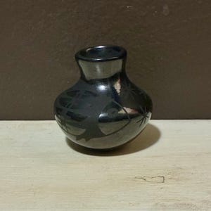 May include: A small, black ceramic vase with a rounded body and a narrow neck. The vase features etched designs, possibly of geometric or floral patterns. The vase sits on a light-coloured wooden surface.