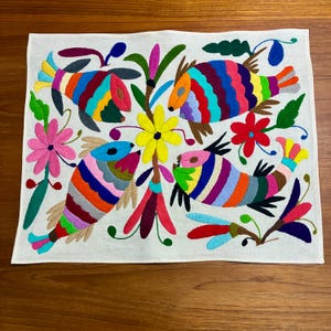 May include: Embroidered white fabric featuring colorful fish and floral designs. The fish are depicted with vibrant stripes in shades of blue, green, red, and yellow. Flowers in pink, yellow, and red add to the decorative appeal. A handcrafted textile.