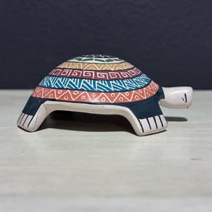 May include: A ceramic turtle figurine with a colorful, geometric pattern on its shell. The shell features bands of orange, blue, green, and brown designs. The turtle's body is white with black accents and a small head.