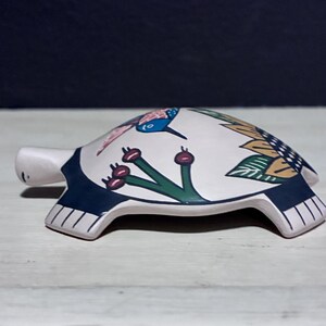 May include: A ceramic turtle figurine with a white shell decorated with a hummingbird, flowers, and leaves. The turtle's head and feet are white, and the shell has a dark blue border. The figurine is on a white surface.