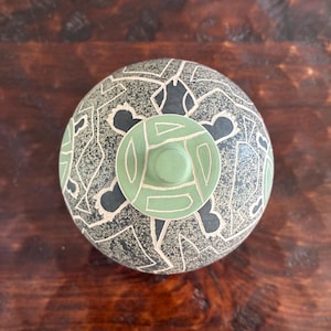 May include: A round, lidded ceramic vessel featuring a dark charcoal grey base with a speckled texture and white geometric line patterns. The lid is topped with a circular, light sage green knob decorated with white carved geometric shapes.