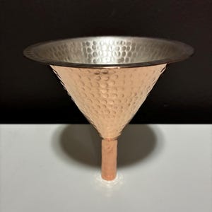 May include: A conical funnel featuring a hammered copper exterior and a smooth silver-toned interior. The wide, flared rim transitions into a narrow cylindrical stem, with the textured metal surface reflecting light across the entire object.