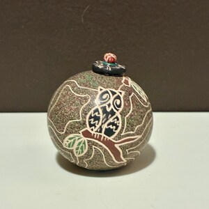 Op de afbeelding: Een bolvormige keramische pot met een getextureerd, gespikkeld bruin oppervlak met een geëtste uil op een tak. Het ontwerp bevat witte, groene en bruine lijnen. Een klein, donker, rond deksel met een rood-witte bloemknop zit bovenop.