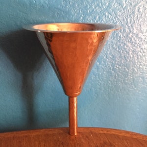 May include: A copper-colored funnel with a hammered texture. The funnel has a wide, flat rim and a long, narrow stem. The funnel is sitting on a wooden surface against a blue wall.