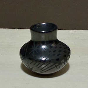 May include: A small, black ceramic vase with a rounded body and a narrow neck. The vase has etched geometric patterns, including a starburst design and a crosshatch pattern, adding visual interest to the dark surface.