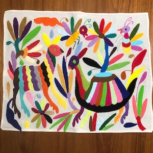 Puede incluir: Textil bordado con un diseño colorido de animales y flores estilizados. La obra de arte incluye un ciervo a rayas arcoíris, un pájaro con cola en forma de abanico y varios motivos florales e insectos, todo ello representado con hilos vibrantes sobre un fondo blanco.