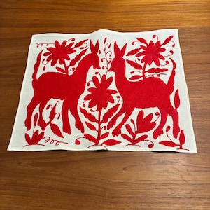May include: White rectangular textile with a red embroidered design featuring two deer and floral elements. The deer are stylized with long legs and upright ears, surrounded by large flowers and leafy stems. The embroidery is detailed and vibrant.