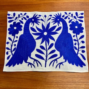 Puede incluir: Textil rectangular blanco con un diseño bordado en azul. El diseño presenta dos aves estilizadas enfrentadas, con una flor grande y elementos florales entre ellas. Las aves y las flores son de un color azul vibrante.
