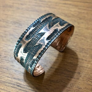 Op de afbeelding: Een koperen manchetarmband met een gedetailleerd, geëtst ontwerp. De armband heeft een reeks parallelle lijnen en driehoekige vormen, met een donkere patina in de geëtste gebieden. De randen zijn afgezet met kleine, verhoogde stippen.