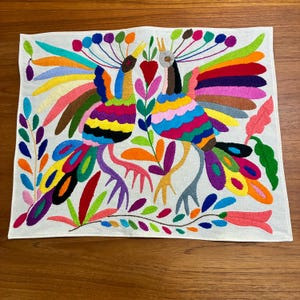 May include: Embroidered textile featuring two vibrantly colored birds with rainbow-hued wings and tail feathers. The birds are set against a white background, surrounded by colorful floral and leaf motifs. The artwork is a lively display of color and design.