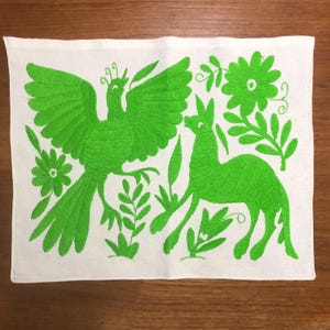 May include: White rectangular textile with vibrant green embroidery. The design features a bird with outstretched wings, a llama-like animal, flowers, and foliage. The embroidery is detailed and the color is bright.