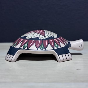 May include: A ceramic turtle figurine with a white base and a shell decorated with a floral pattern in burgundy, green, and white. The turtle's shell has a dark blue border. The figurine has a smiling face.