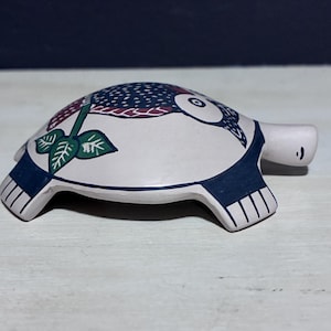 May include: A ceramic turtle figurine with a white body, navy blue accents, and a detailed shell design. The shell features a pattern of navy blue dots, red and white accents, and a green leaf design. The turtle has a small, white head.