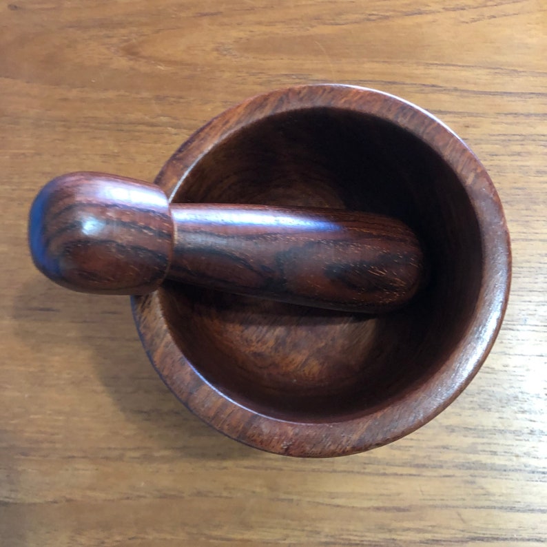 May include: A wooden mortar and pestle set. The mortar is a round bowl with a dark brown finish. The pestle is cylindrical with a rounded end, also in a dark brown wood. The set is designed for grinding herbs and spices.