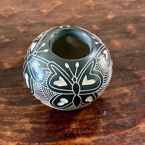 May include: A dark green, round decorative vase with a carved butterfly design. The butterflies are white and outlined with intricate patterns. The vase has a circular opening at the top.