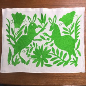 May include: White rectangular textile with embroidered green rabbits and floral designs. The rabbits are depicted in a playful pose amidst the foliage. The embroidery is detailed, creating a textured appearance. The textile is likely a decorative item.