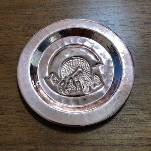 May include: A silver-colored metal coaster with a hammered finish. The coaster features a raised design of a mountain range with a crescent moon above it.