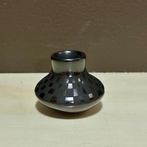 May include: A small, dark grey ceramic vase with a wide, rounded base and a narrow neck. The vase features a geometric pattern of dark squares across its body. The interior and rim are a solid, darker grey.