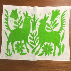May include: White rectangular textile with vibrant green embroidered design. Features two stylized deer figures amidst floral and foliate motifs. The embroidery is detailed, creating a textured appearance. The textile is likely a decorative piece or a kitchen towel.