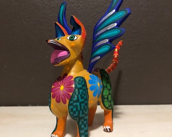 May include: A colorful wooden figurine of a dog with wings. The dog is painted in shades of yellow, orange, blue, green, and pink. The wings are blue and silver. The dog has a wide open mouth and a playful expression.