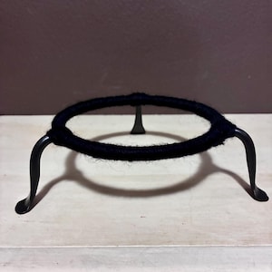 May include: A black metal stand with a circular top wrapped in dark blue yarn. The stand has three curved legs and a central support. The object is designed to hold a round object, such as a bowl or a decorative item.