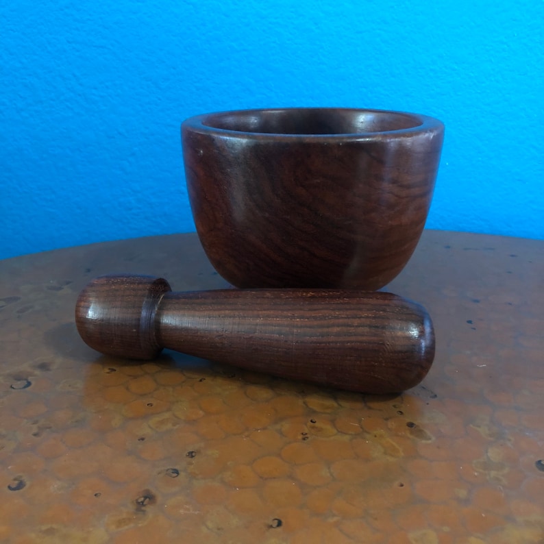May include: A dark brown wooden mortar and pestle set. The mortar is a bowl shape, and the pestle is cylindrical with a rounded handle. The set is made of a dark wood with visible grain.
