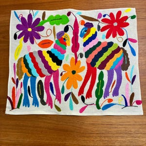 May include: Embroidered textile featuring two stylized, colorful animals with striped bodies and floral accents. The animals are depicted in vibrant rainbow hues, surrounded by flowers and foliage in shades of purple, red, orange, and green on a cream background.