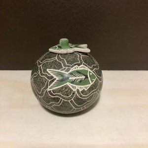 Op de afbeelding: Een ronde, groen en grijze decoratieve pot met een visontwerp. De pot heeft een deksel met een visvormige handgreep. Het visontwerp is groen en wit en de pot heeft een getextureerd oppervlak.