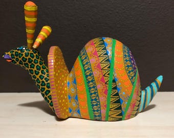 May include: A hand-painted wooden snail figurine with vibrant colors and intricate patterns. The snail's body features orange, green, blue, and pink geometric designs. The head has a leopard print pattern, and the antennae are striped.