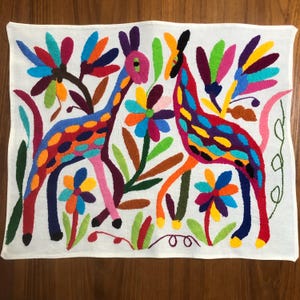 May include: Embroidered textile with two giraffes and floral designs. The giraffes are in blue and red with colourful spots, surrounded by flowers in orange, purple, and green. The background is white. The textile is a vibrant piece of folk art.