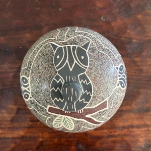 May include: A round, speckled grey stone lid featuring a central dark grey owl carving perched on a brown branch. The owl has white etched details, and additional stylised owl faces are visible on the sides of the circular stone surface.